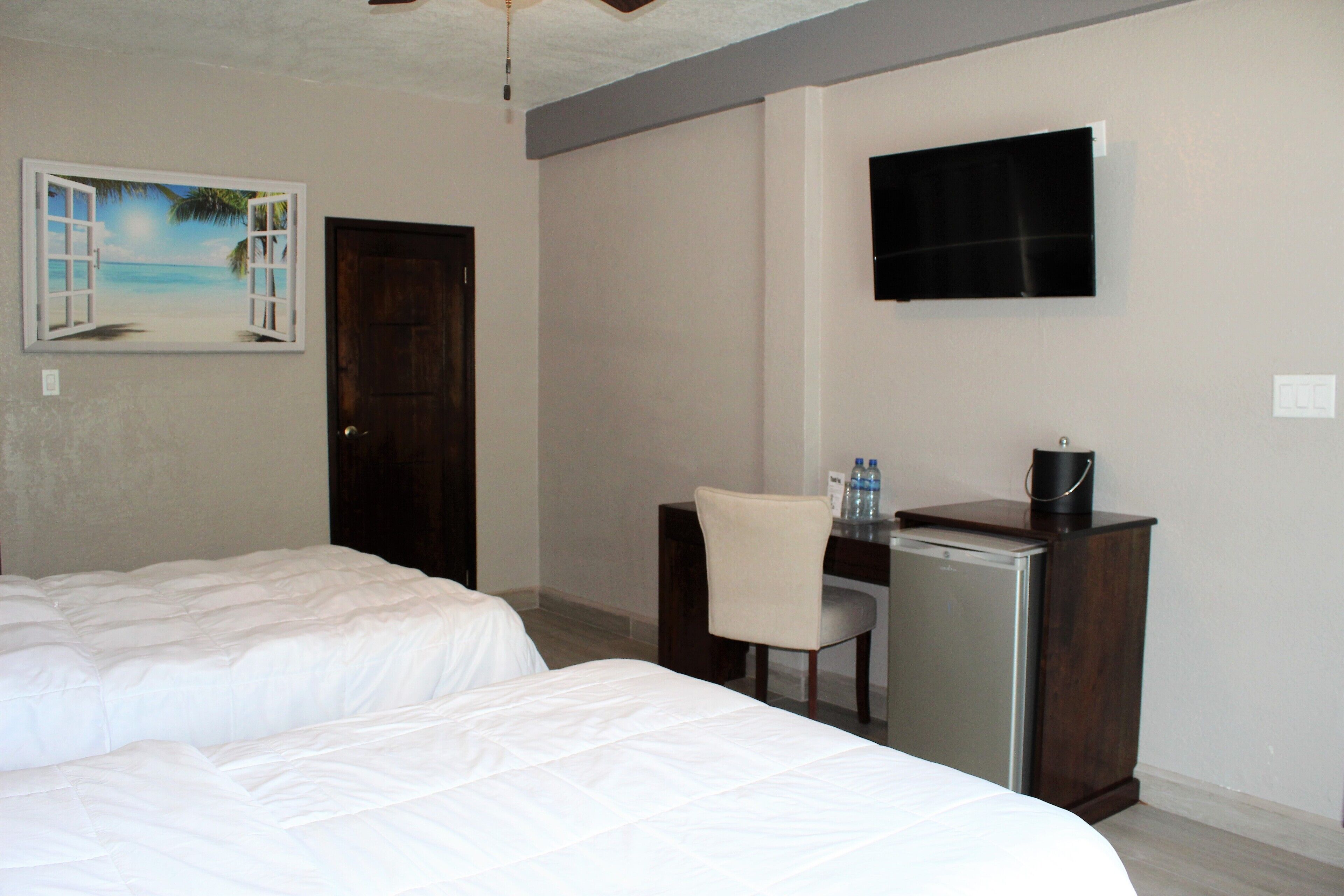 A hotel room with two beds, a desk with a chair, a television, and a painting of a beach scene.