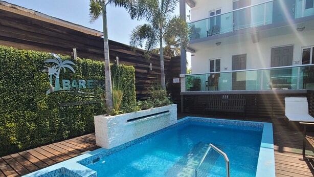 A swimming pool with a wooden deck, surrounded by greenery and a building with a balcony.