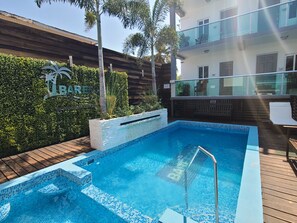 A swimming pool with a wooden deck, surrounded by greenery and a building with a balcony. - Barefoot Caye Caulker Hotel (Caye Caulker)