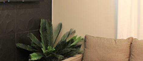 Un salon moderne avec un téléviseur à écran plat fixé au mur, un canapé beige, un fauteuil jaune et une grande plante en pot.