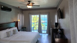 A hotel room with a bed, ceiling fan, air conditioning unit, and a view of the ocean through large windows.
