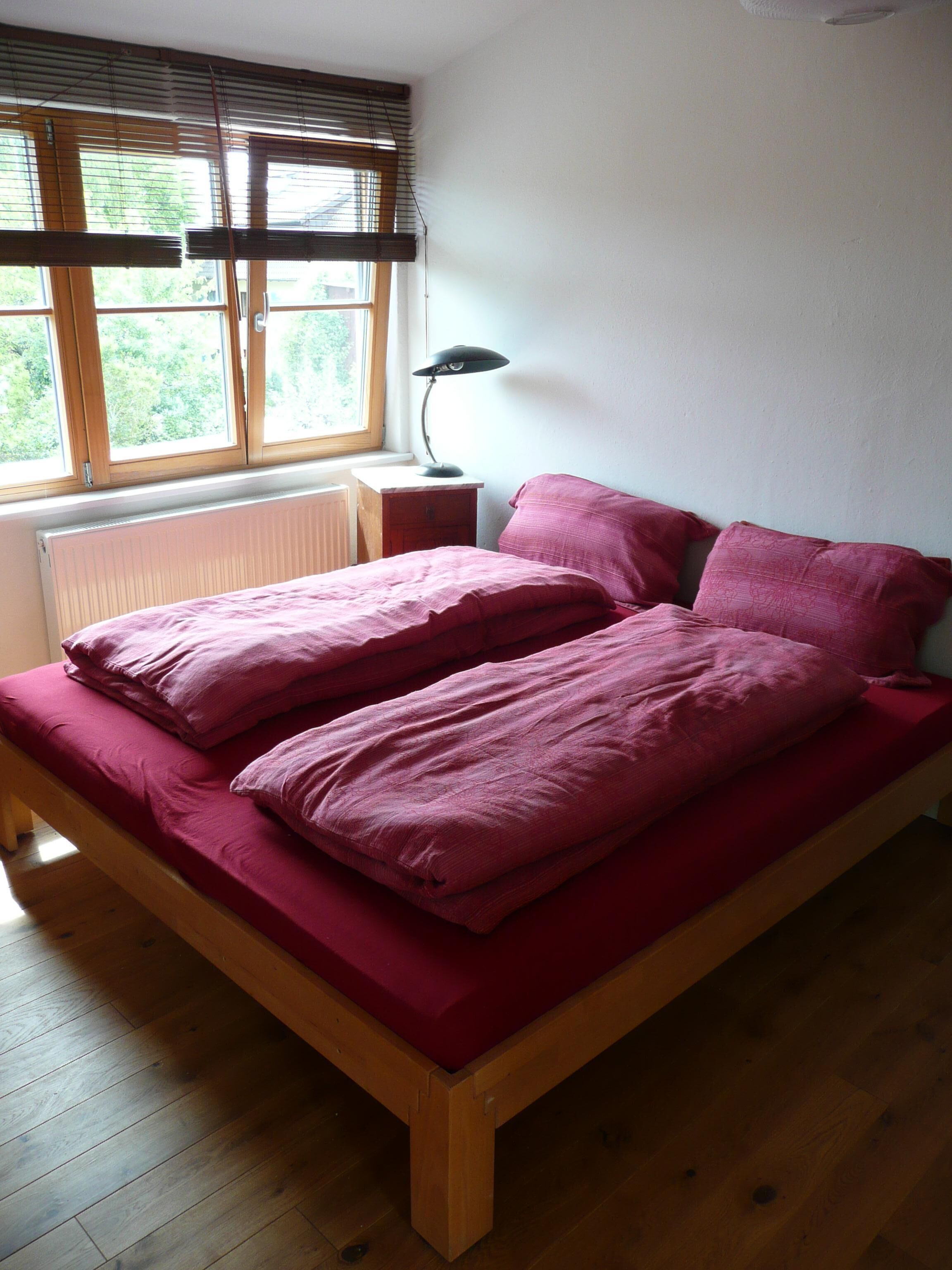 A single bed with a wooden frame and a red mattress, a bedside table with a lamp, and a window with blinds.