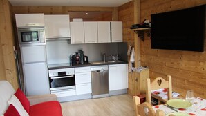 A compact kitchen with white cabinets, a microwave, an oven, and a dishwasher. There is a dining area with a table set for two, a red and white sofa, and a wooden chair with a heart-shaped backrest. - Renovated 2-bedroom apartment, station center, WIFI (La Clusaz)