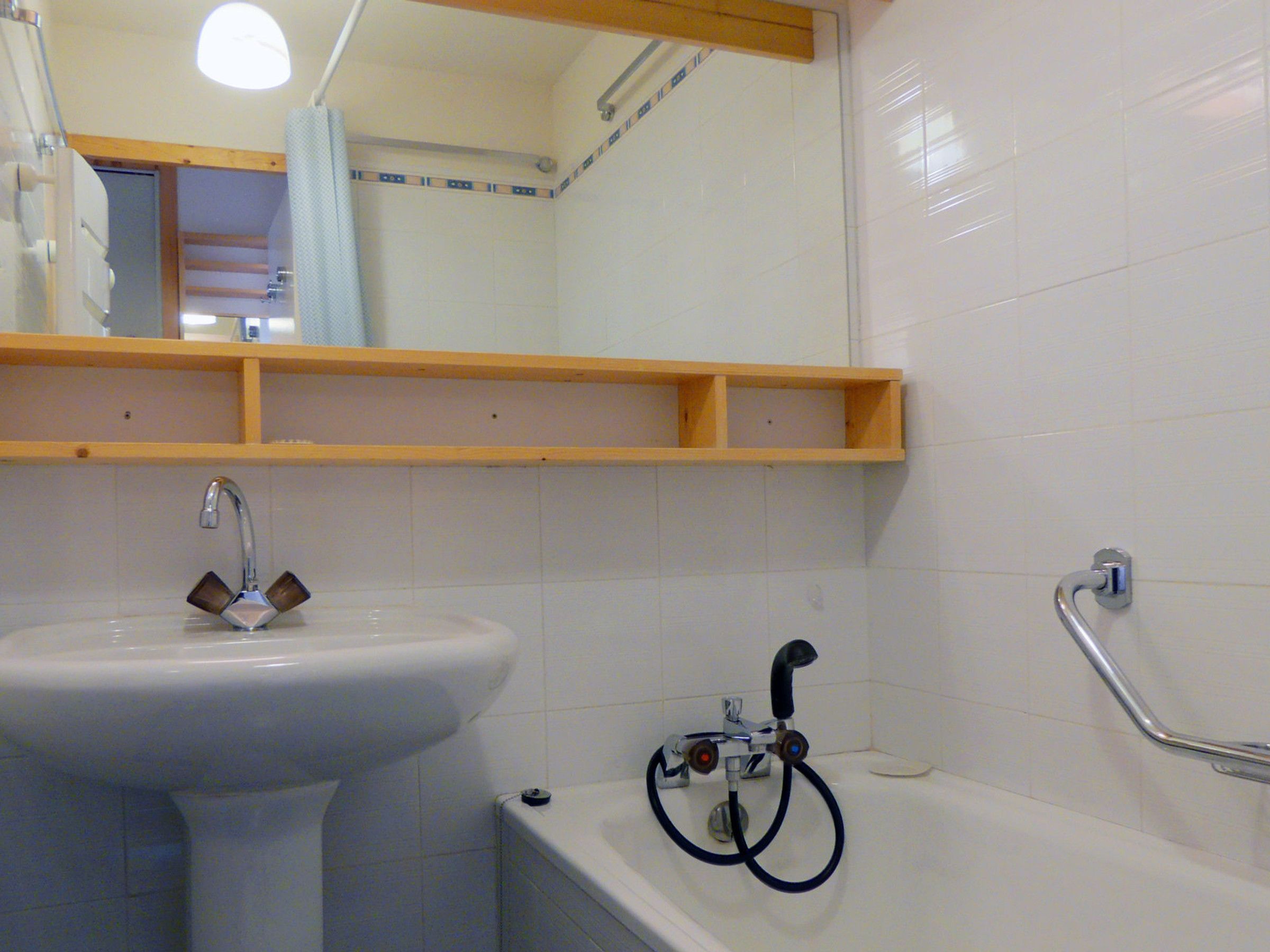 A bathroom with a white sink, a bathtub, and a shower. There is a wooden shelf above the sink and a handrail on the wall next to the bathtub.