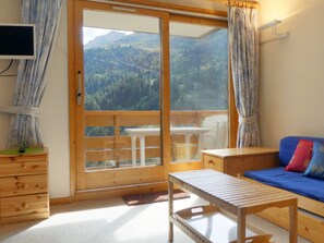 A room with a wooden bench, a blue sofa with colorful cushions, a wooden table, and a view of a forested mountain through a glass door. - Family apartment 6 pax, south balcony, near Méribel (Méribel)