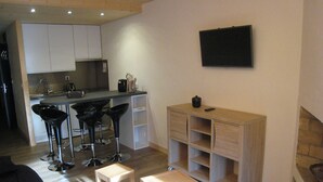 A compact living space with a kitchenette, a dining area with bar stools, a TV mounted on the wall, and a small wooden cabinet. - 2-Room Apartment, Ski-In/Ski-Out, South Balcony, Comfort for 6 (La Clusaz)