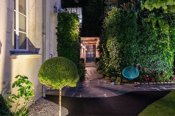 A well-maintained garden pathway leading to a building entrance, surrounded by lush greenery and a small seating area.