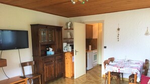 A cozy living room with a wooden floor, a TV mounted on the wall, a dining table set with a tablecloth, and a kitchen in the background. - 1-bedroom near slopes with balcony (La Clusaz)