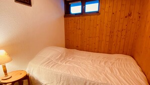 A bedroom with a wooden headboard, a single bed with a white duvet, a small wooden stool, and a wall-mounted lamp. - Cozy 2-Room Apartment with Balcony and Mountain View in La Clusaz (La Clusaz)