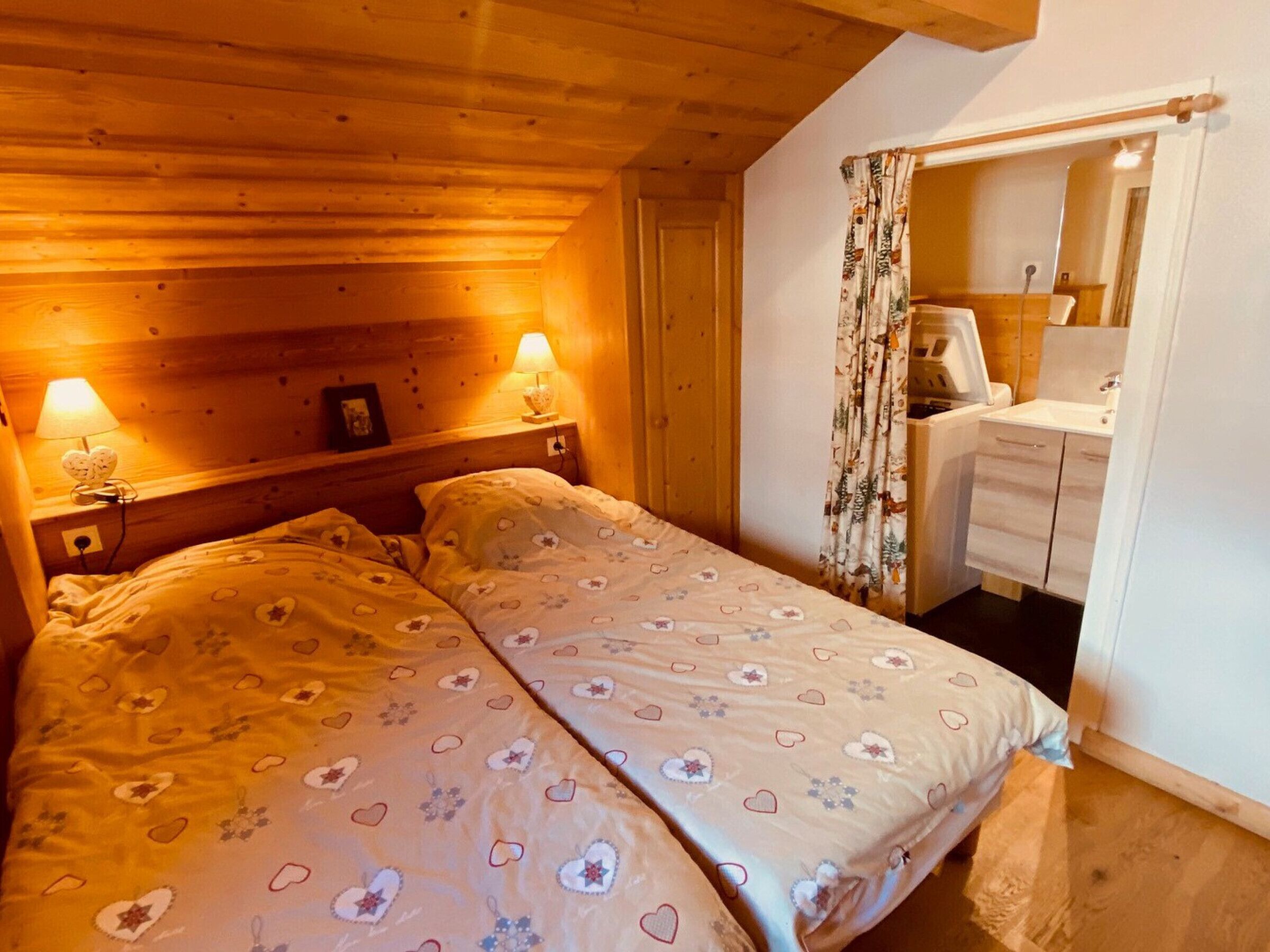 A cozy bedroom with a wooden ceiling, a double bed with heart-patterned bedding, bedside lamps, and a bathroom with a mirror and sink.