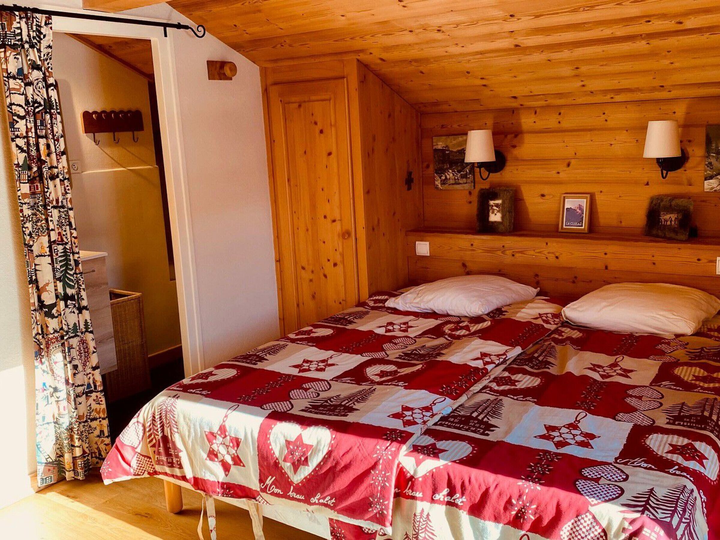 A cozy room with a wooden ceiling and walls, a bed with a red and white quilt, two wall-mounted lamps, and a small shelf with decorative items.