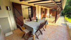 A covered patio with a table and chairs, wooden doors, and a view of the garden.