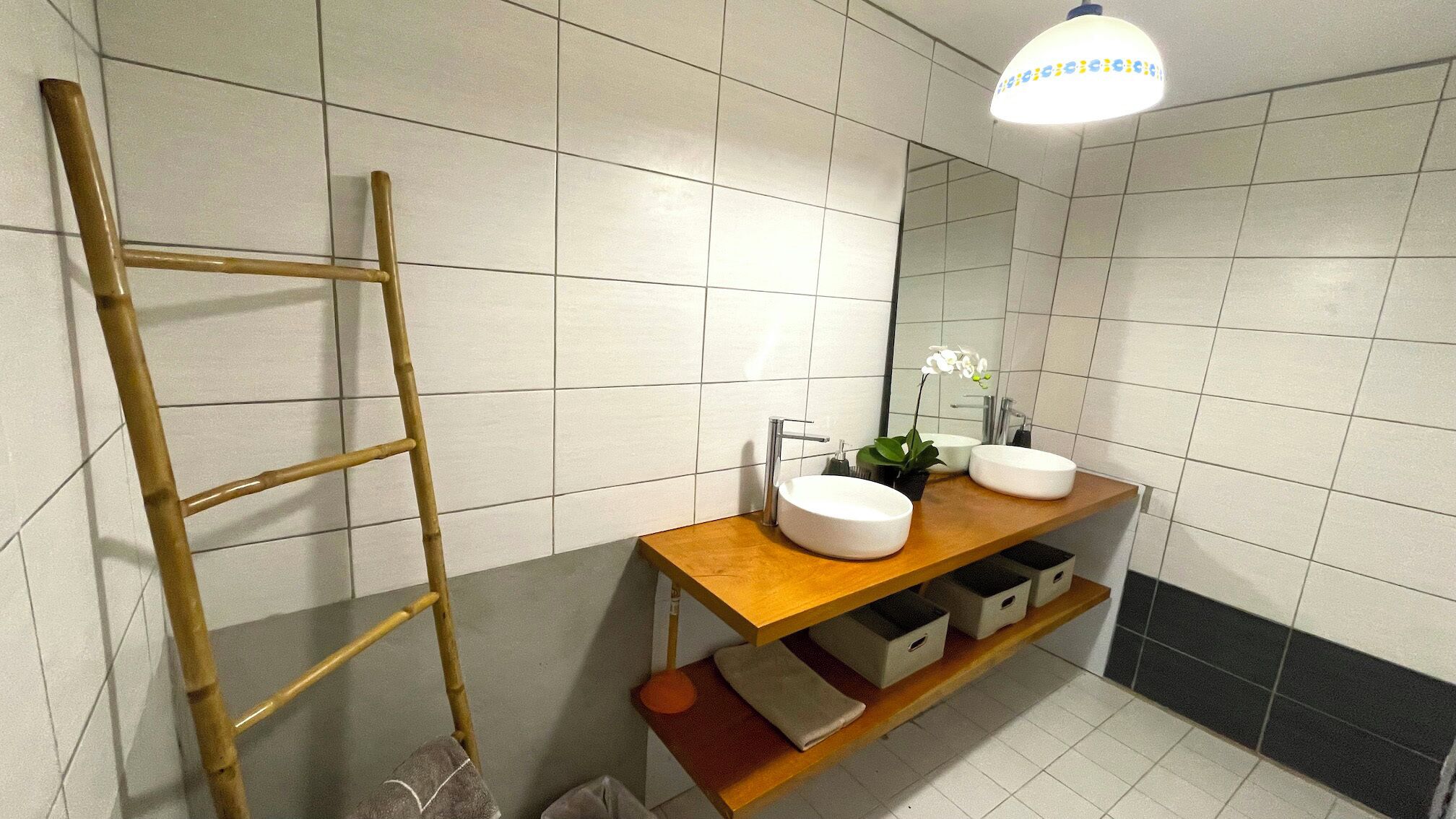 A bathroom with white tiled walls, a wooden vanity with two sinks, a mirror, a potted orchid, and a wooden ladder.