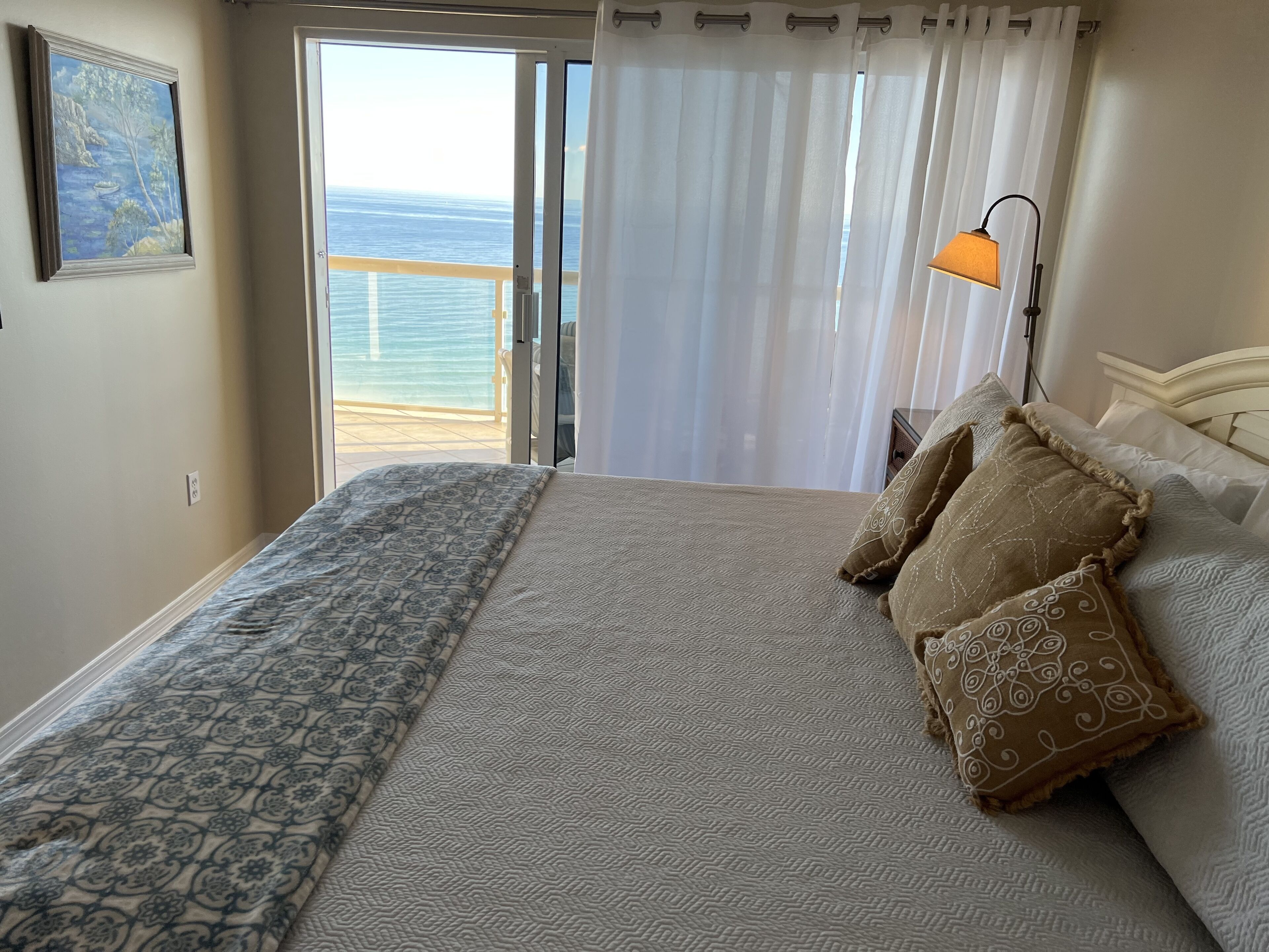 Een slaapkamer met een groot bed, uitzicht op de oceaan door een schuifdeur en een lampje op het nachtkastje.