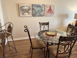A dining area with a glass table surrounded by wicker chairs, a metal chair, and a decorative plate. Three artworks depicting a crab, a starfish, and an octopus are displayed on the wall. - 𝔾𝕦𝕝𝕗𝔽𝕣𝕠𝕟𝕥 𝑬𝒎𝒆𝒓𝒂𝒍𝒅 𝑰𝒔𝒍𝒆 9th Floor Stocked & Stellar Reviews! (Unknown)