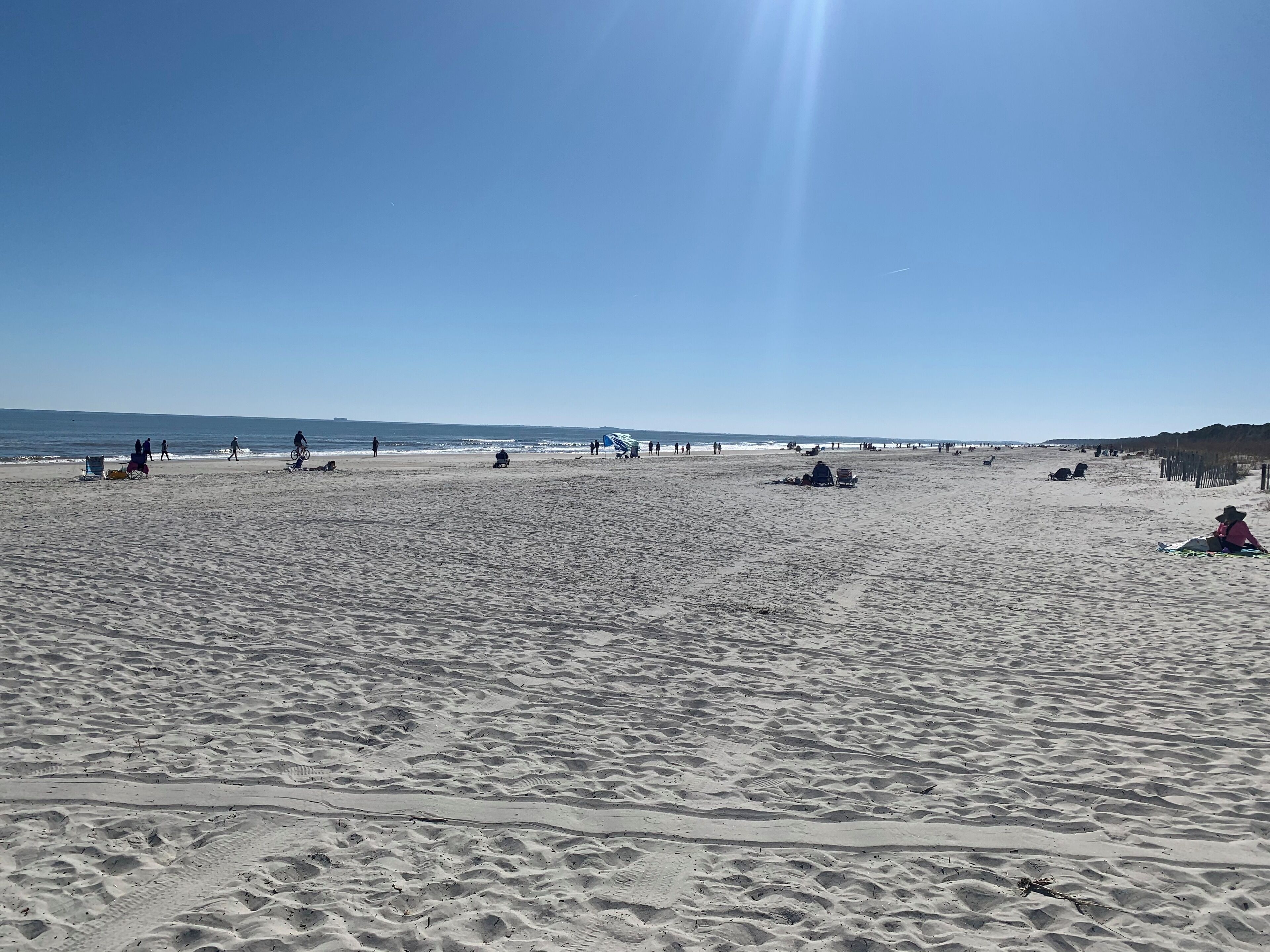 A wide sandy beach with people walking and sitting, a clear sky, and a distant pier.