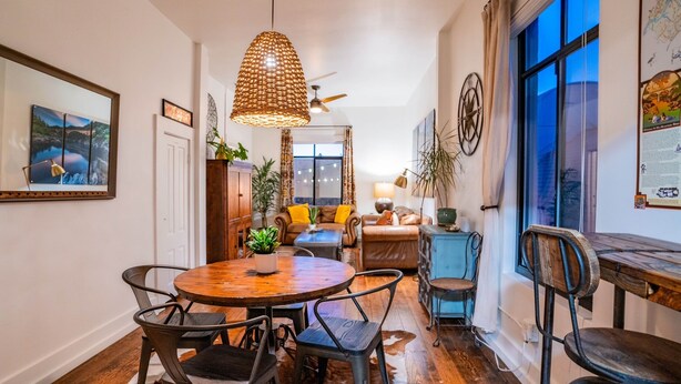 A dining area with a round wooden table and metal chairs, a hanging woven light fixture, and a view of a living room with a sofa and cushions.