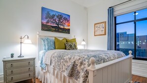 A bedroom with a bed, bedside tables, a nightstand, a window with a view, and a painting on the wall.