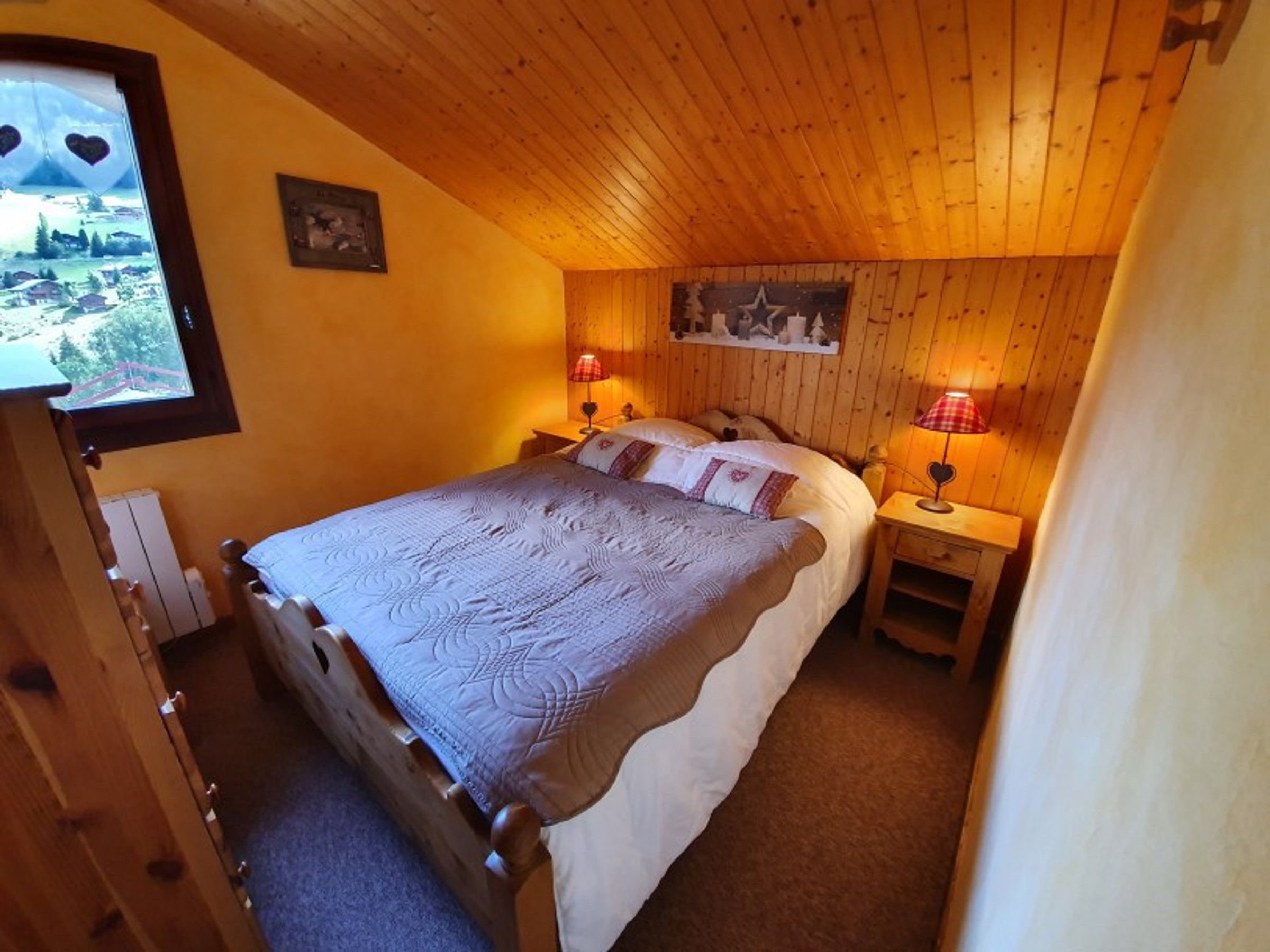 A bedroom with a wooden ceiling, a bed with a quilt, two bedside tables with lamps, and a window with a view of a landscape.