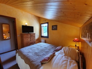 Une chambre avec un plafond en bois, un lit recouvert d’une courtepointe, une commode, un téléviseur, une fenêtre donnant sur un beau paysage et une lampe fixée au mur.