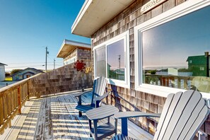 A wooden deck with Adirondack chairs, a large window, and a view of the ocean.