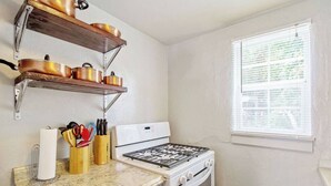 A kitchen with a stove, oven, and shelves holding pots. There is a window with blinds and a countertop with a paper towel holder and utensils. - Affordable for Long Stay, Great for Budget-Friendly Business Travels (Oklahoma City)