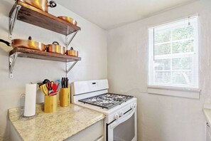 Una cucina con fornello, forno e mensole piene di pentole. C'è una finestra con persiane e un piano di lavoro con porta asciugamani di carta e utensili.