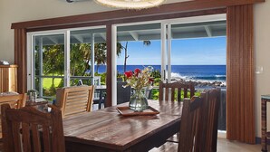 Un comedor con mesa de madera, sillas y un florero. Amplias ventanas ofrecen vista al océano y la playa.