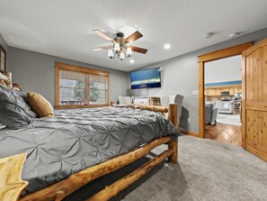 A bedroom with a bed, wooden headboard, a ceiling fan, a TV, and a view of the kitchen through an open door.