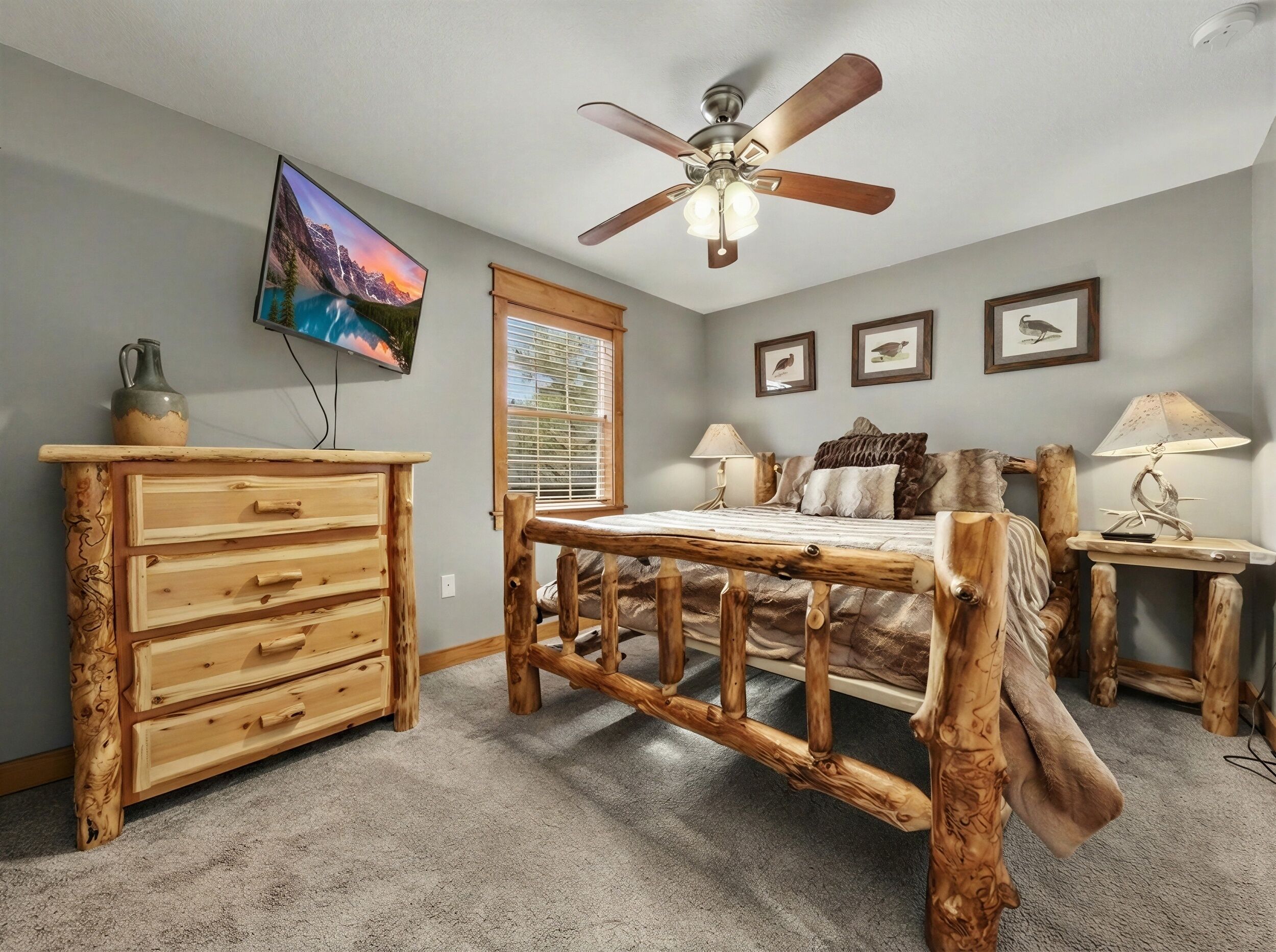 A bedroom with a wooden bed frame, a ceiling fan, a TV mounted on the wall, and a window with blinds.