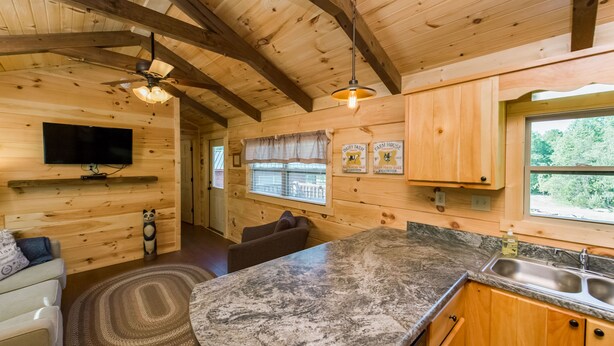 Interior de una cabaña de madera con una zona de cocina, una mesa de comedor y un televisor montado en la pared.