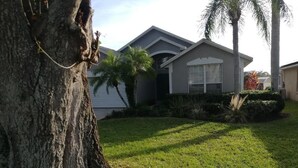 A residential house with a well-maintained lawn and a large tree in the foreground. - Villa Bonita. Southern Dunes 4 star golf course. Disney and Legoland area. (Haines City)