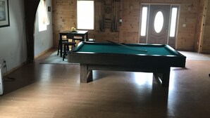 A pool table in a room with wooden walls and a sloped ceiling. There is a table with chairs and a window with curtains. - Bring your boats, jetskis, and LARGE family - so much space inside and out (Sylvan Beach)