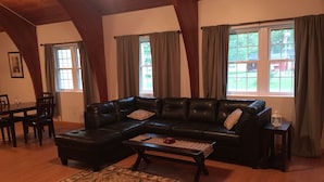 A living room with a black leather sectional sofa, a wooden coffee table, and a patterned rug. There are two windows with curtains, a dining table with chairs, and a small side table with a lantern. - Bring your boats, jetskis, and LARGE family - so much space inside and out (Sylvan Beach)