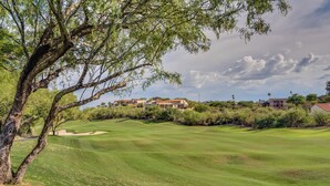 A golf course with a large tree in the foreground, houses in the background, and a clear sky.