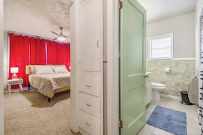 Een slaapkamer met een bed, nachtkastje, kledingkast, plafondventilator en een badkamer met toilet en wastafel.