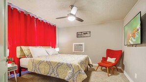 A bedroom with a bed, a red chair, a ceiling fan, and a wall-mounted TV.