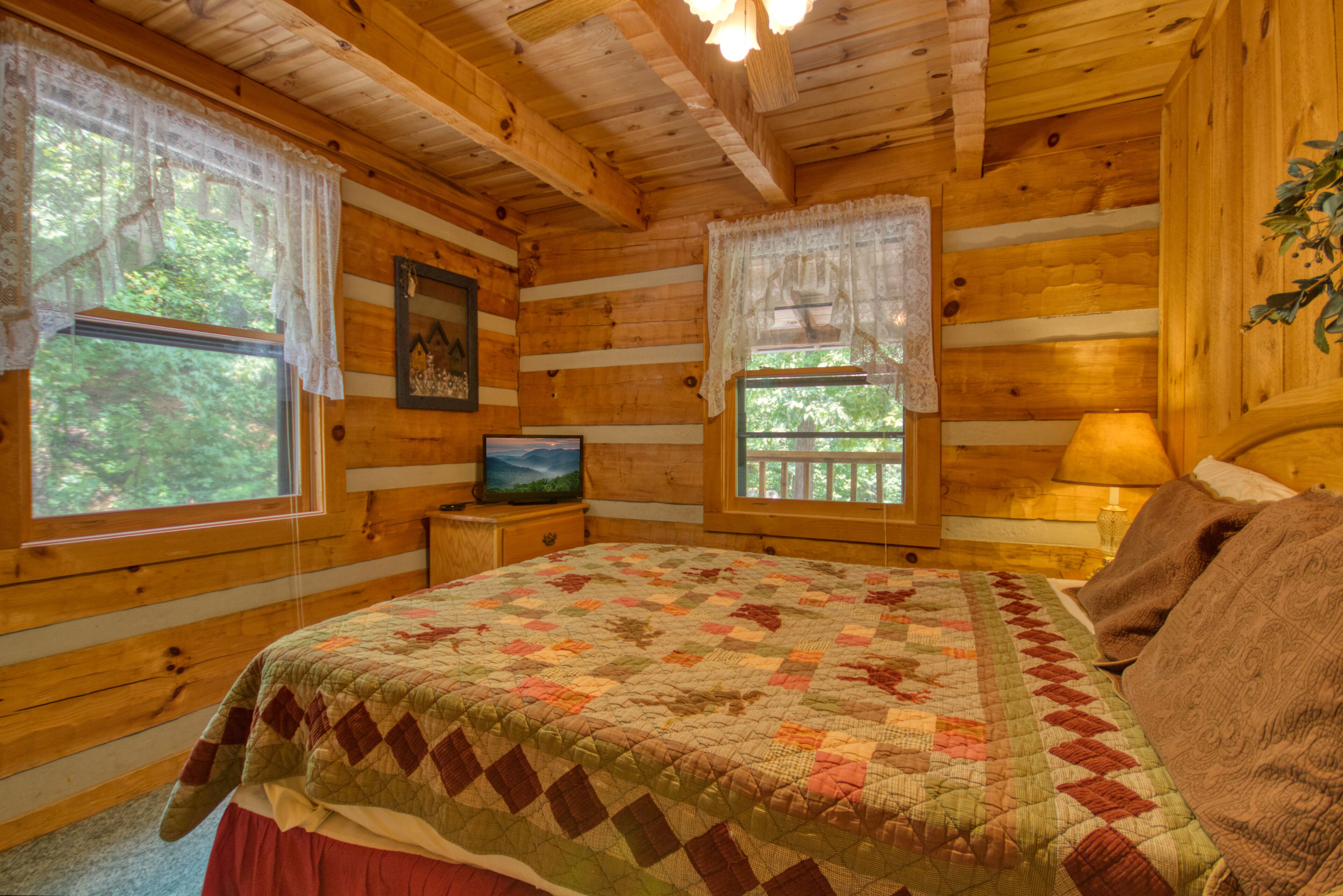 Une chambre confortable dans un chalet en rondins, avec une couvre-lit matelassé, un téléviseur posé sur une table de chevet et une fenêtre donnant sur la forêt.