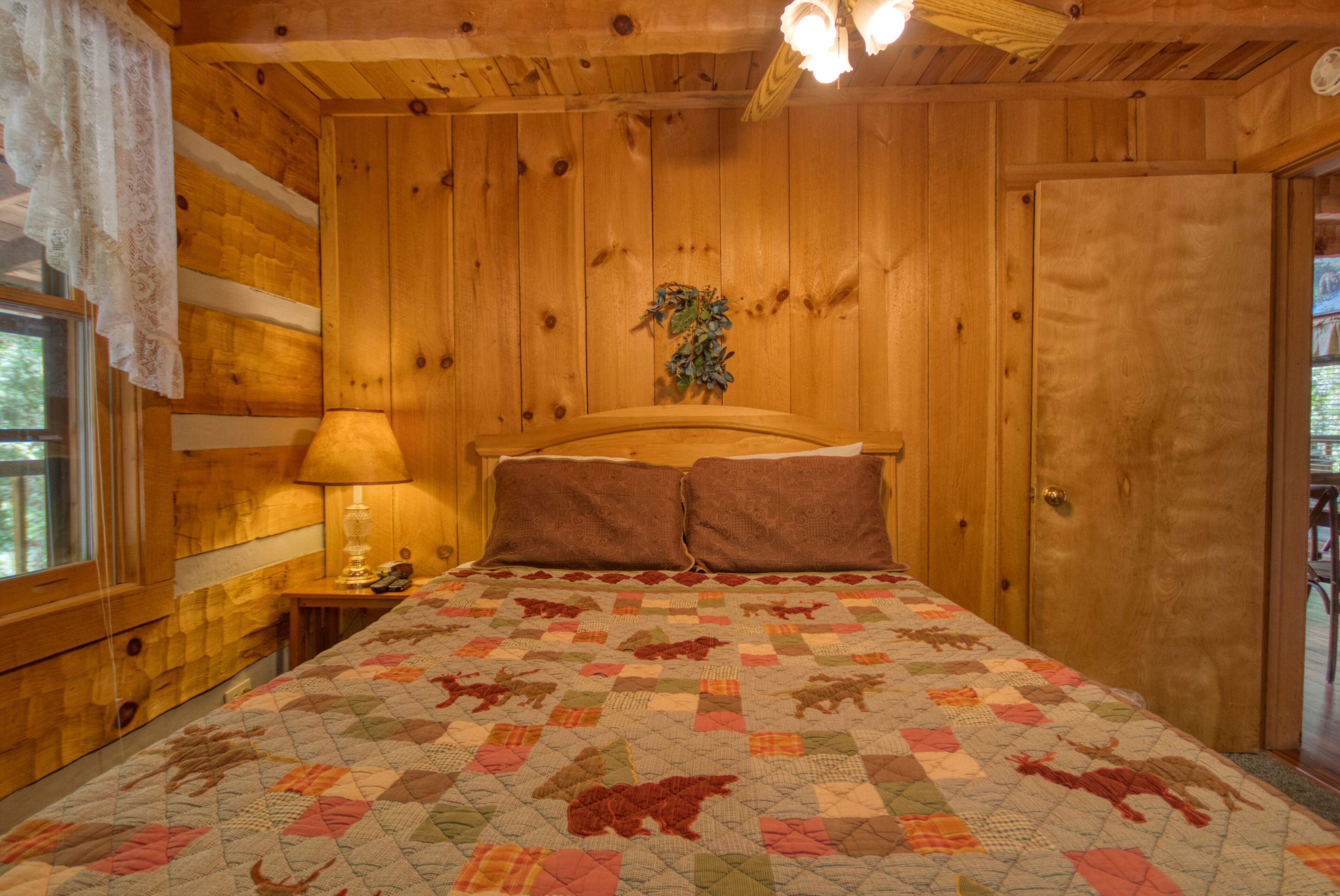 A wooden cabin bedroom with a quilted bedspread, a nightstand with a lamp, and a door to the right.