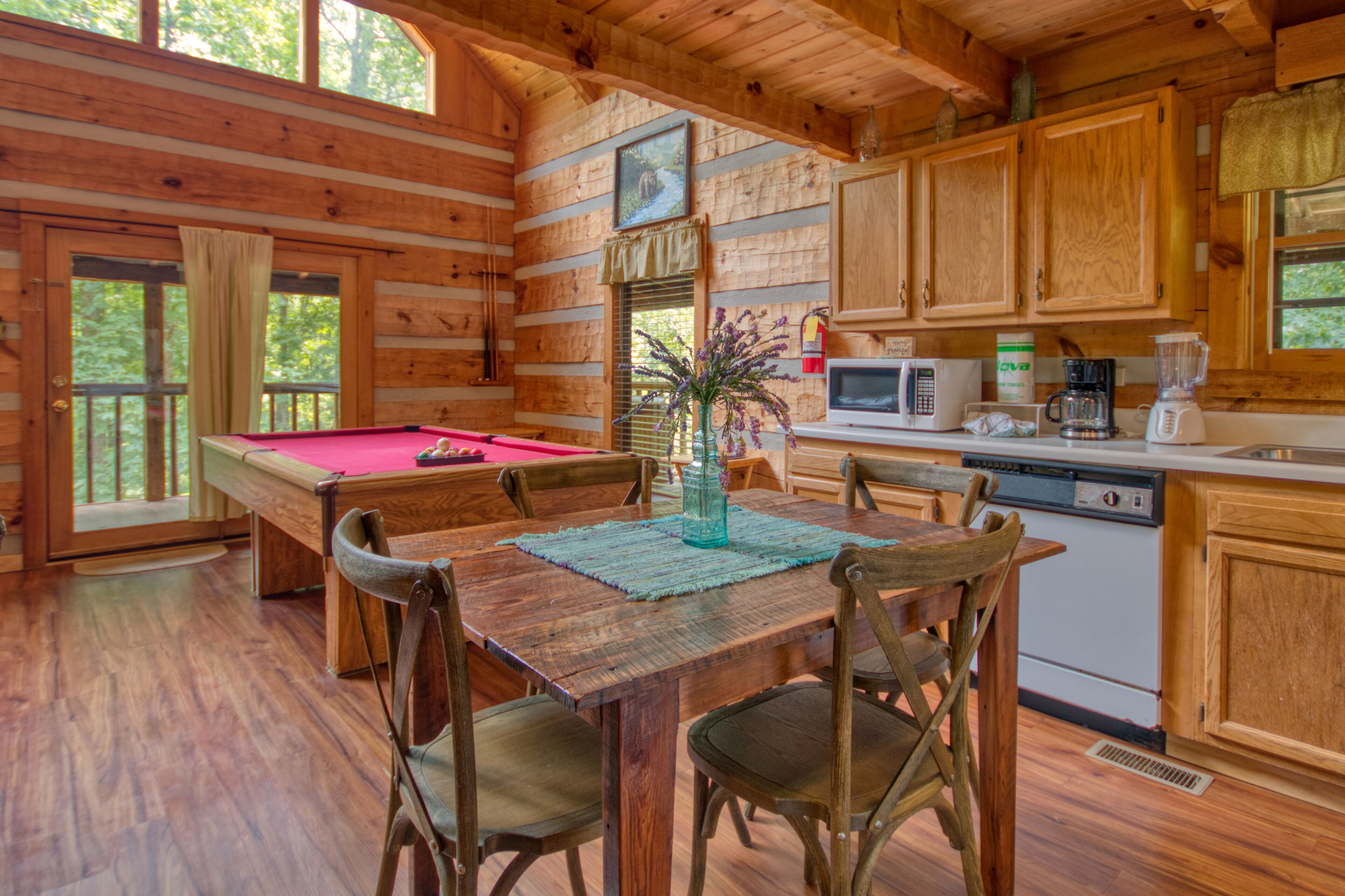 A wooden cabin interior with a pool table, dining table, chairs, kitchen area, and wooden cabinets.