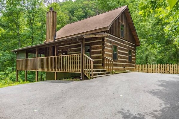 A rustic log cabin with a porch and a chimney, surrounded by dense forest.