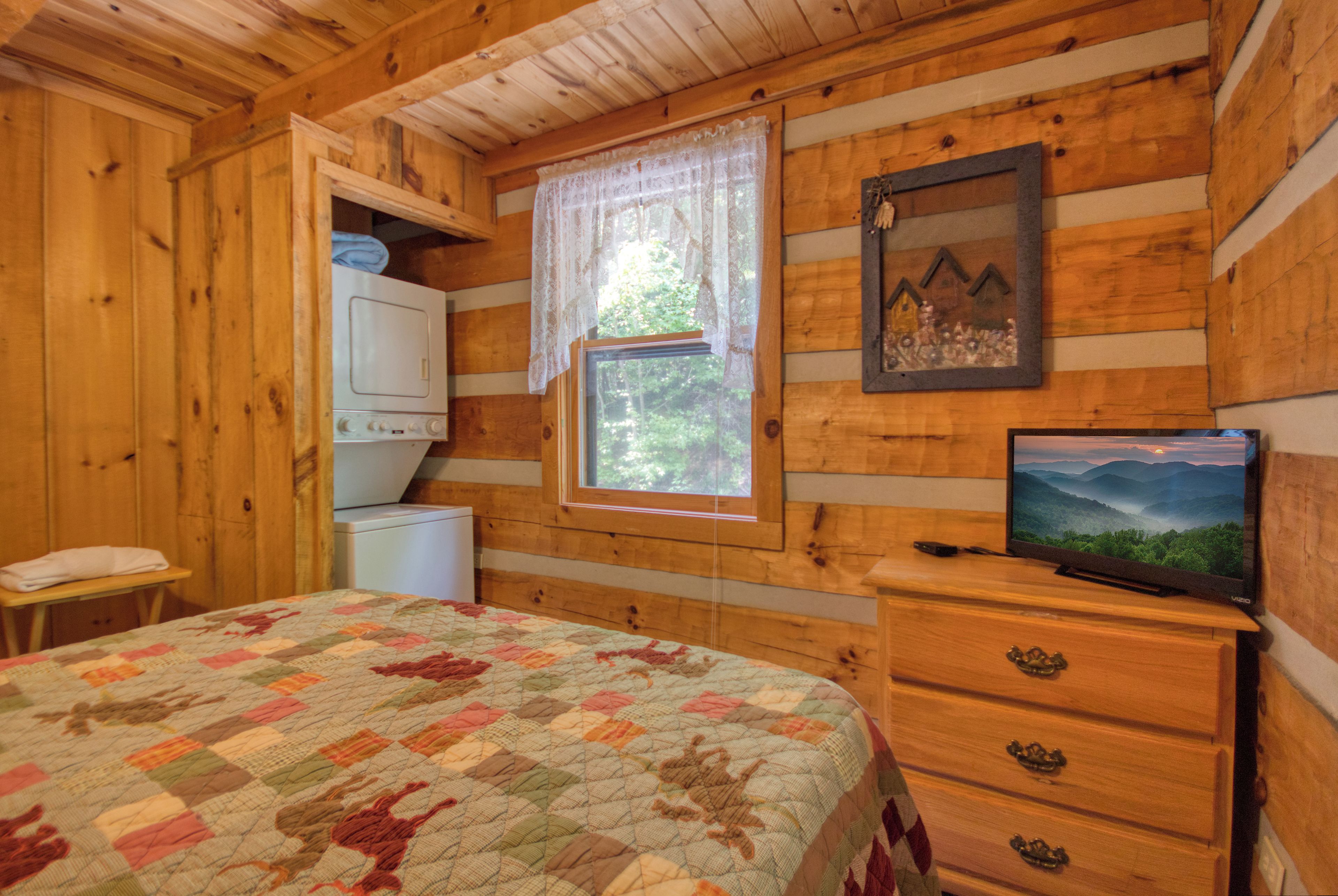 A wooden cabin bedroom with a quilted bed, a television on a dresser, and a window with a view of greenery.
