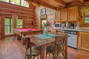 A wooden cabin interior with a pool table, dining table, chairs, kitchen area, and wooden cabinets.