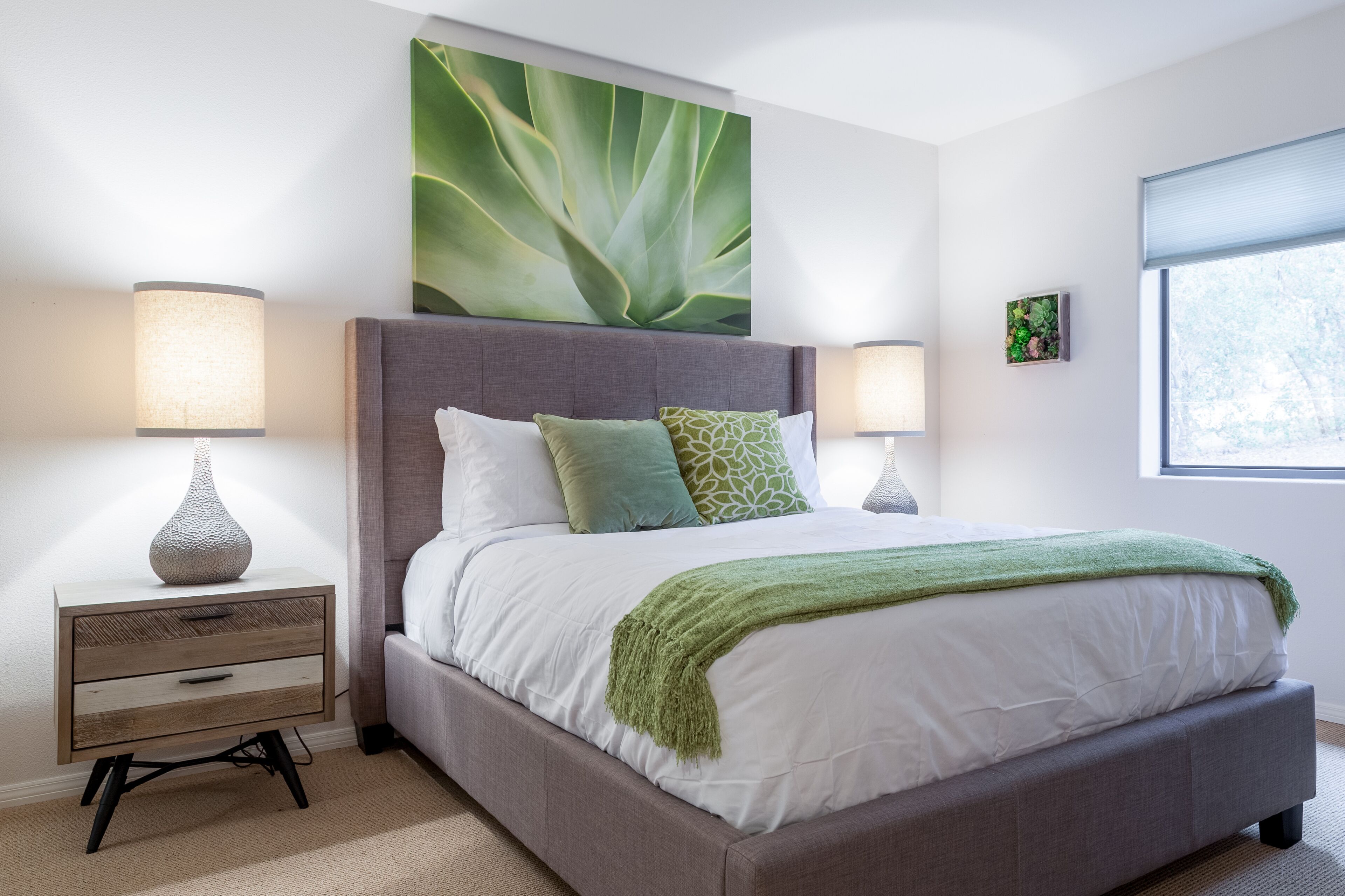 Un dormitorio moderno con una cama tapizada en gris, ropa de cama blanca y detalles en verde. Hay dos mesitas de noche con lámparas, una gran obra de arte verde en la pared sobre la cama y una ventana con persianas a la derecha.