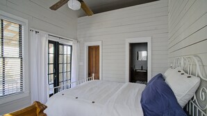 Une chambre à coucher avec un grand lit, une tête de lit en bois et un ventilateur de plafond. Il y a deux portes et une fenêtre avec des stores.