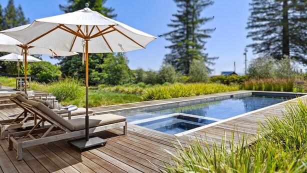 A wooden deck with lounge chairs and umbrellas, a swimming pool, and a natural landscape with trees.