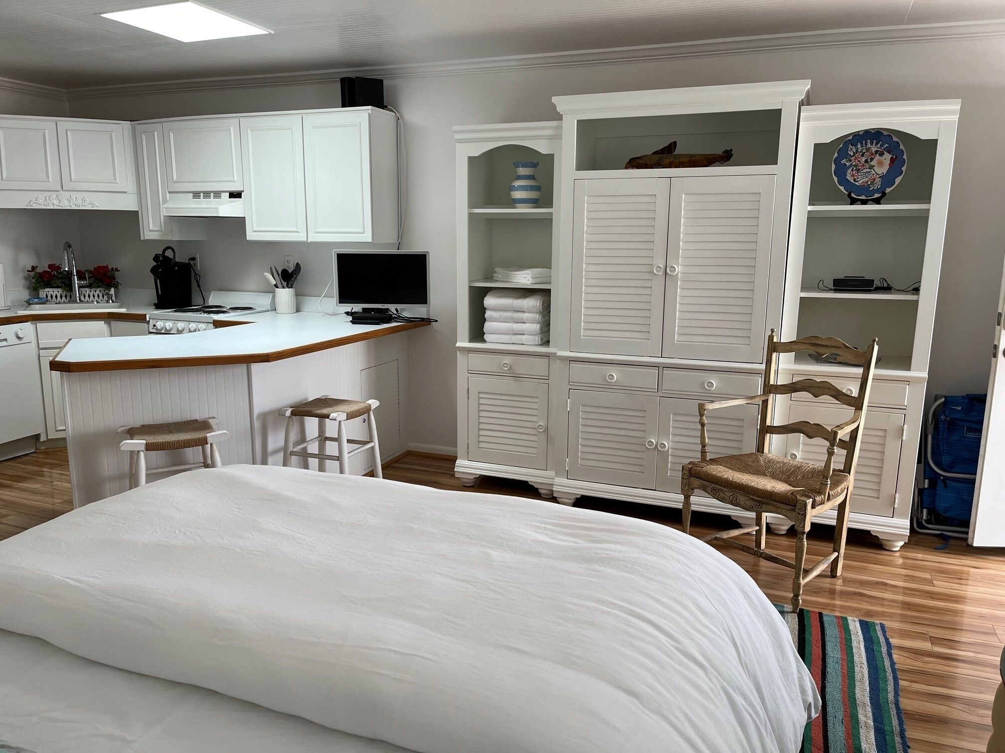 A bedroom with a large bed, a wooden chair, a desk with a computer, and a kitchenette.