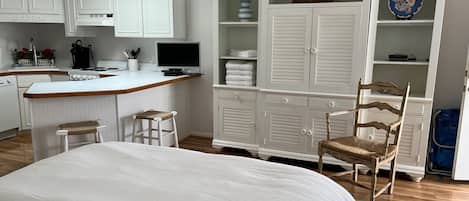 A bedroom with a large bed, a wooden chair, a desk with a computer, and a kitchenette.