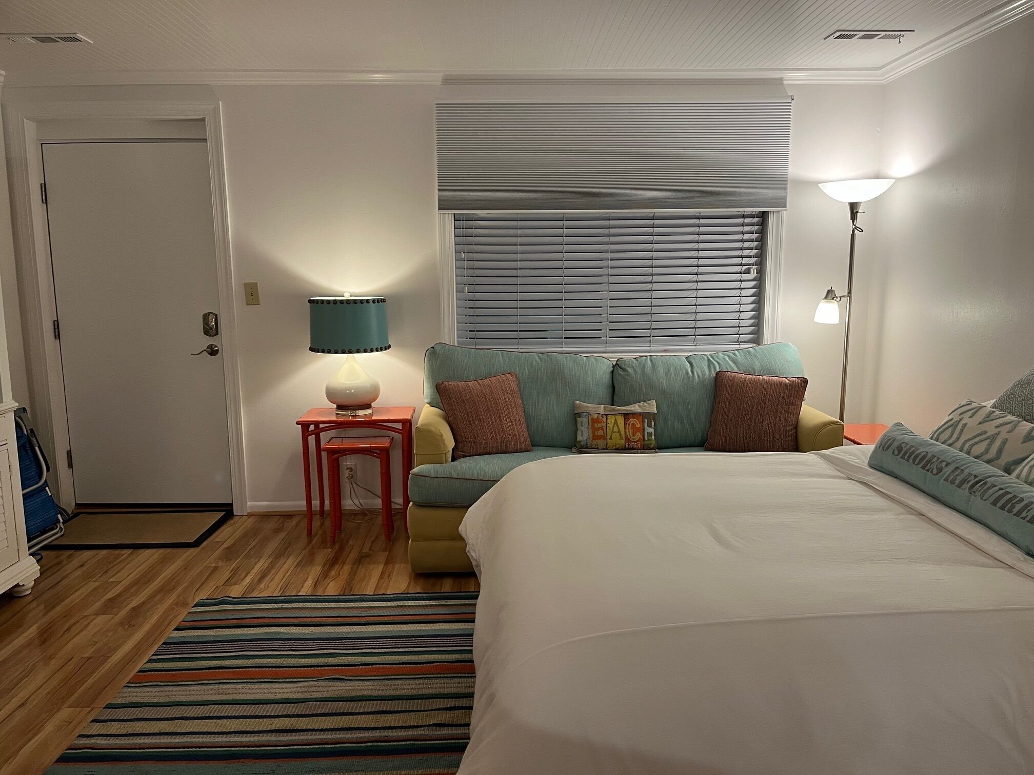 A bedroom with a bed, a sofa, a small table with a lamp, a window with blinds, and a door.