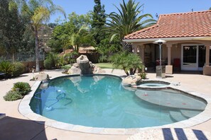 A swimming pool with a rock feature and a small hot tub, surrounded by palm trees and a building with a red-tiled roof. - San Diego home with RESORT BACKYARD!! (Poway)